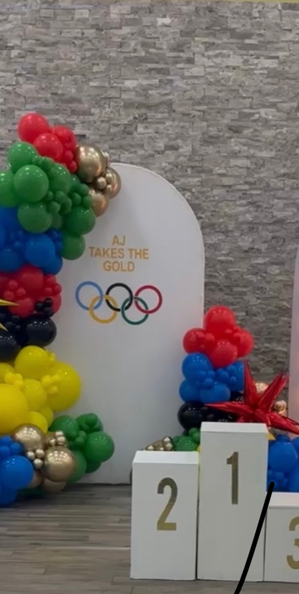 Decorative setup with balloons, Olympic rings, and a sign reading 'AJ Takes the Gold' against a stone wall.